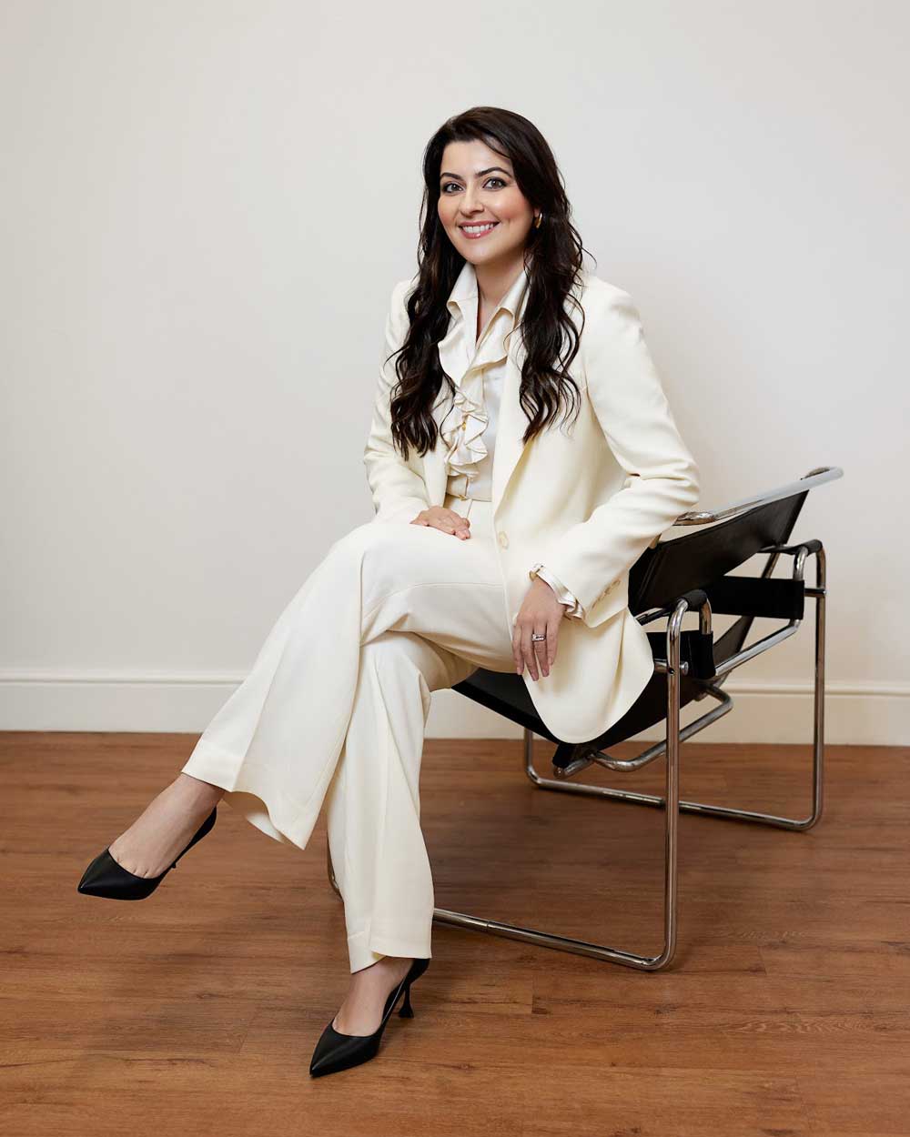 Dr Hawkes seated on a modern black chair, dressed in an elegant white suit with a ruffled blouse, on a wooden floor against a plain beige background.