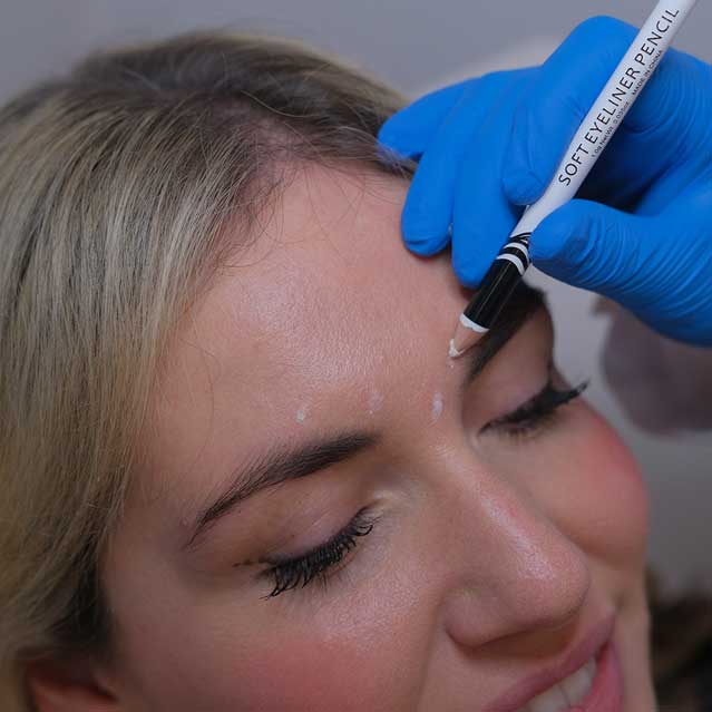 Close-up of a woman having dots marked on her forehead with a soft eyeliner pencil by a professional wearing blue gloves.
