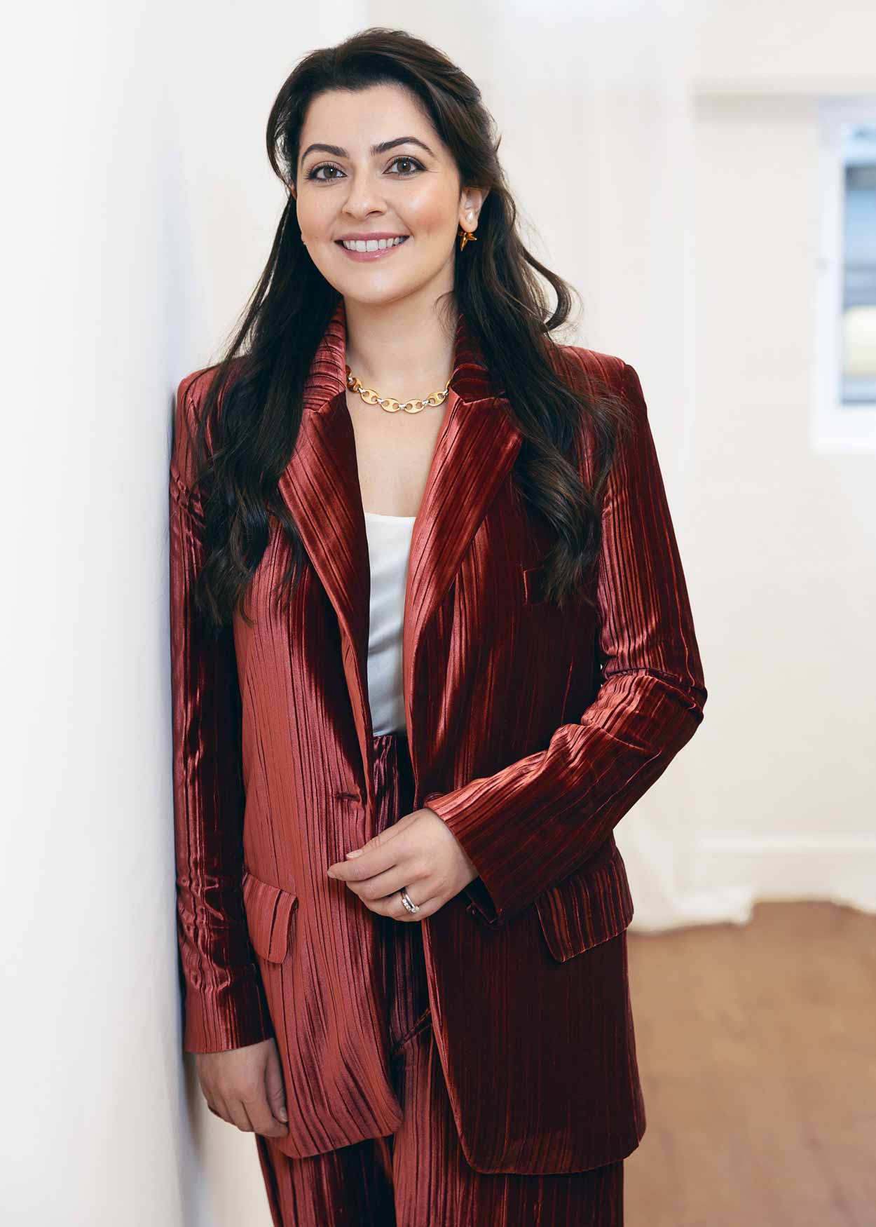 Dr Hawkes wearing a red pleated suit with a white top and gold necklace, leaning against a white wall in a bright room.